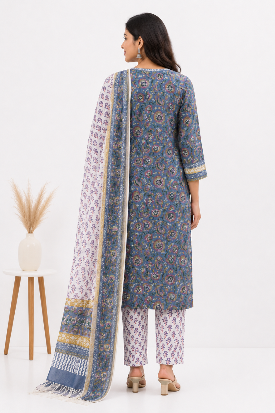 A woman shown from the back wearing a blue patterned kurta with matching printed pants and a long patterned dupatta, photographed in a clean studio setting with a small side table and vase.