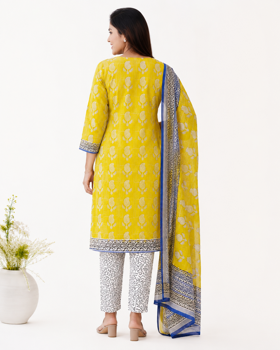 A woman shown from the back wearing a bright yellow traditional outfit with beige floral motifs, patterned pants, and a matching yellow dupatta with blue and borders, photographed against a clean white studio background.