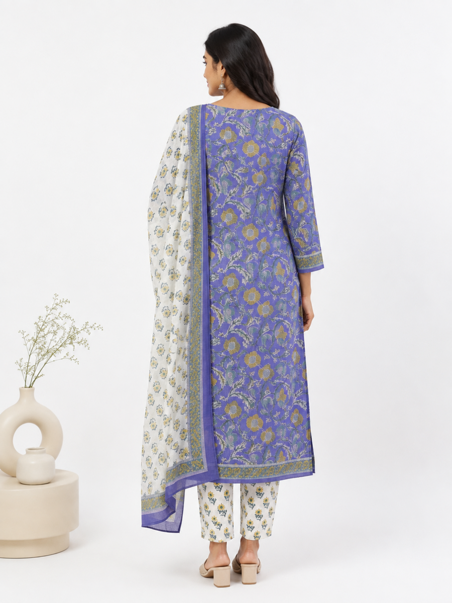 A woman is shown from the back wearing a traditional three-piece salwar kameez set. The set features a royal-blue printed kurta with gold and green floral motifs, matching printed trousers, and a white dupatta with a blue border and gold floral print.