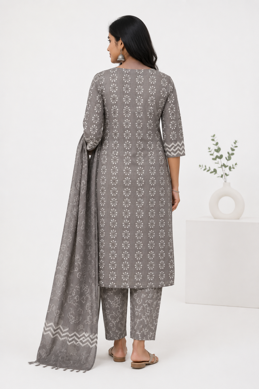 A woman stands with her back to the camera wearing a gray three-piece salwar kameez suit with white floral prints and a matching dupatta. The set includes a long kurta, matching trousers, and a draped dupatta with a chevron pattern at the border 