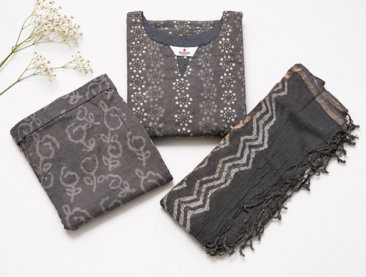 Three folded garments and a matching dupatta set in dark gray and charcoal tones with white floral and geometric patterns, arranged on a white background with small white flowers as styling props.