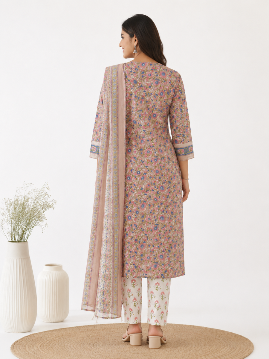 A woman stands with her back traditional three-piece salwar kameez suit. The outfit features a dusty pink tunic with a multicolor floral print, matching patterned trousers, and a coordinating sheer dupatta draped over her shoulder.